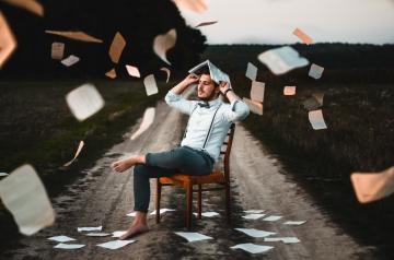 A person sitting on a chair outside on a dirt road, eyes closed, with an open book on the end with pages flying around